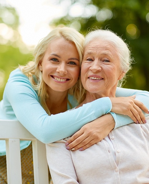 An older woman is sitting on a bench while another woman hugs her and smiles