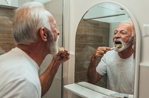 An older man is standing in front of his mirror brushing his teeth in his bathroom